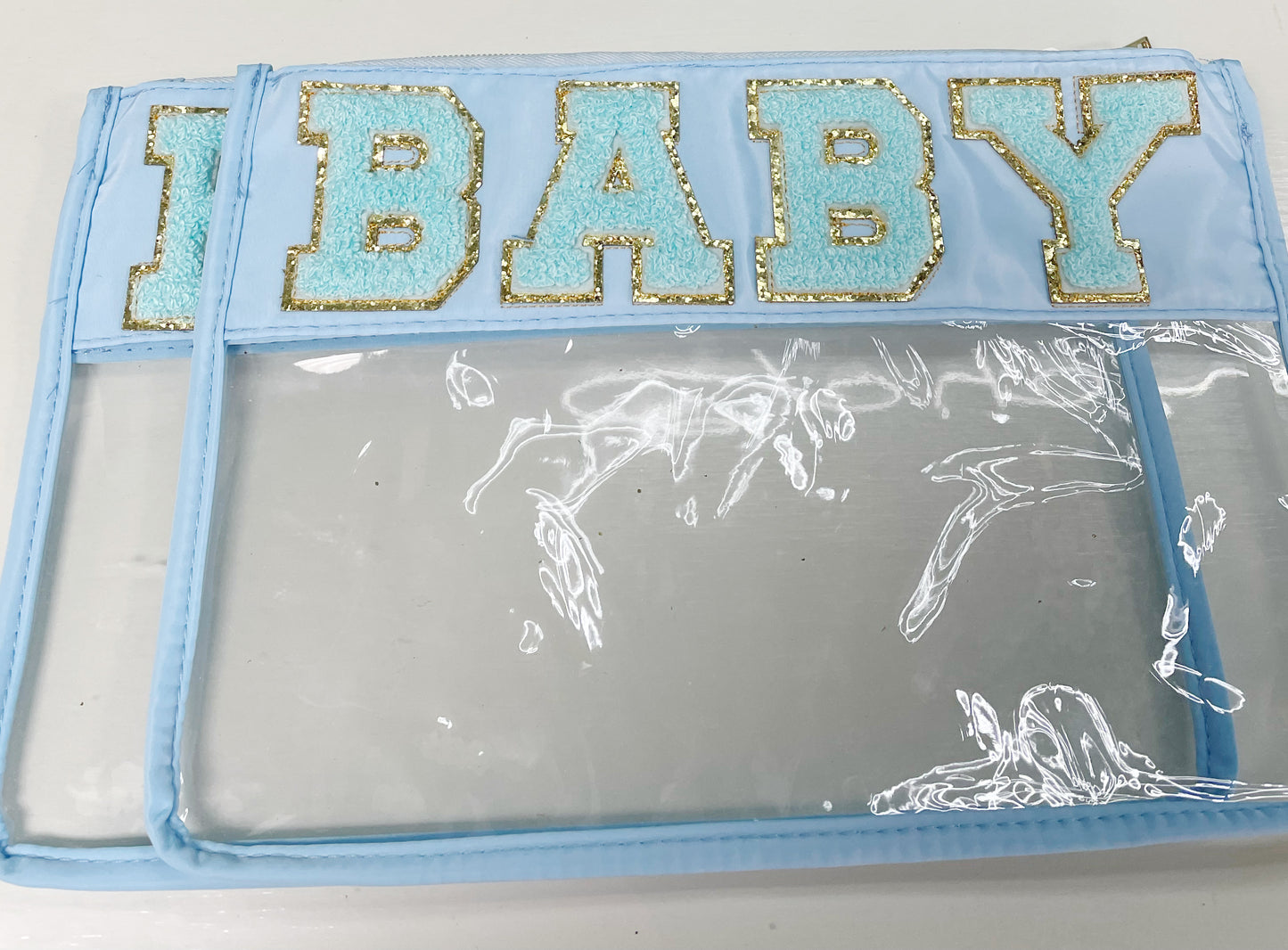 Clear zippered pouch that has a light blue border with the word 'baby' embroidered at the top in chenille light blue varsity letters outlined in shiny gold.