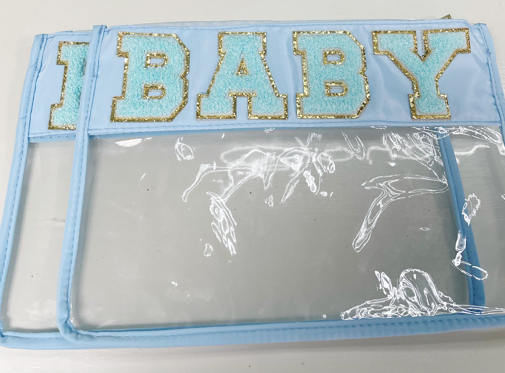 Clear zippered pouch that has a light blue border with the word 'baby' embroidered at the top in chenille light blue varsity letters outlined in shiny gold.